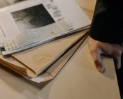 Close-up of a hand resting on a desk near a stack of investigative documents and newspaper clippings.