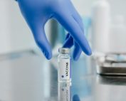 Close-up of a vaccine vial on a metal surface with a gloved hand in a laboratory setting.