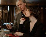 A woman prepared to light a cigarette while a man offers a lighter, set in an elegant bar setting.