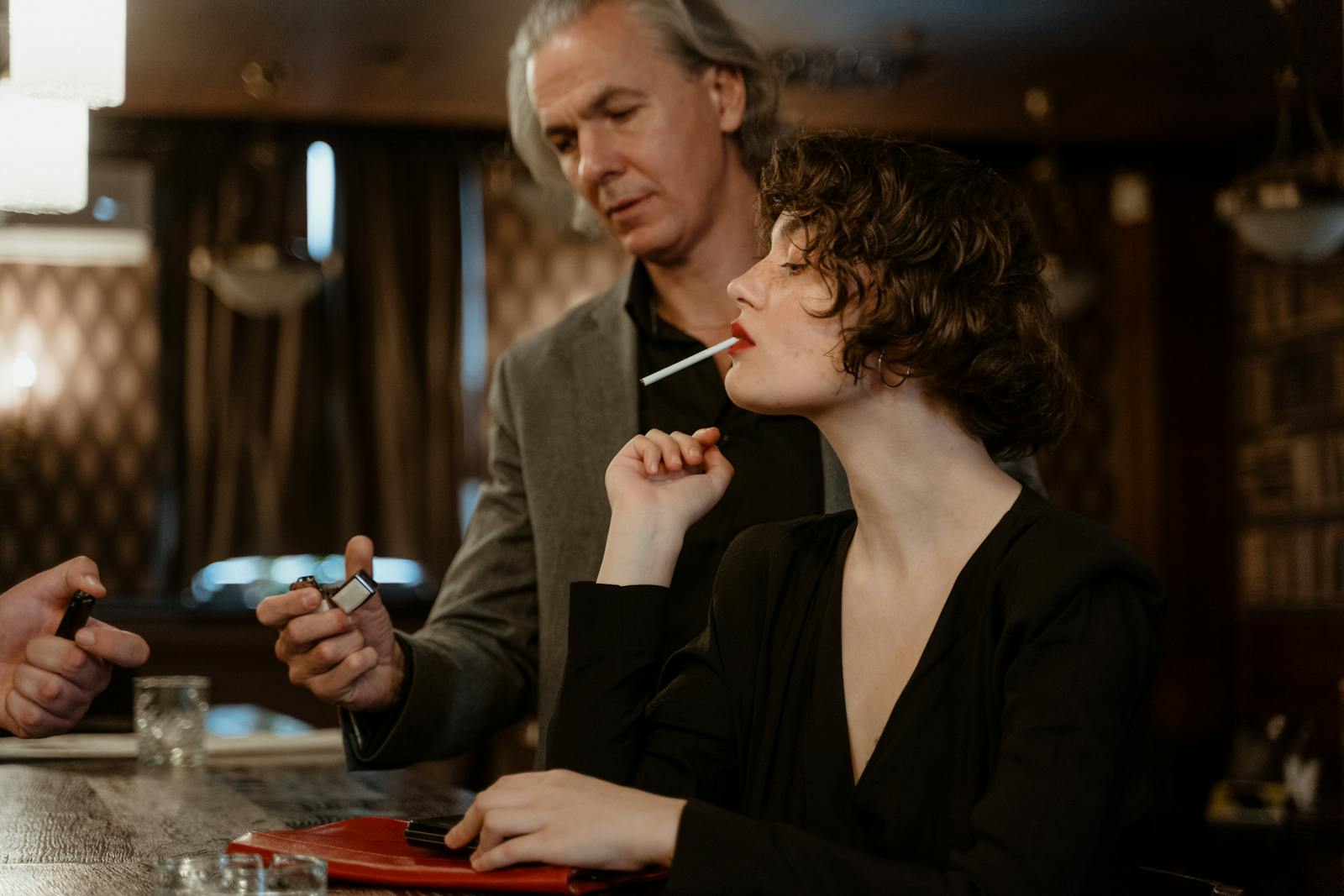 A woman prepared to light a cigarette while a man offers a lighter, set in an elegant bar setting.