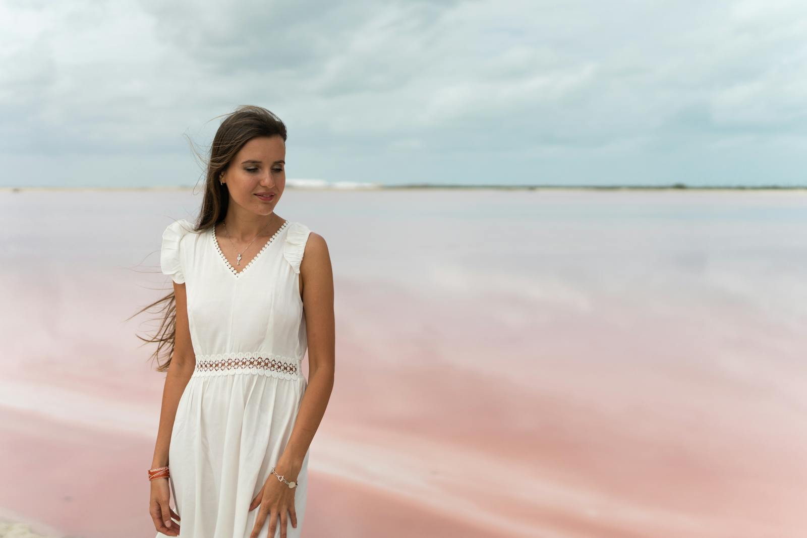 A woman standing by a serene pink lake wearing a white dress, exuding tranquility.