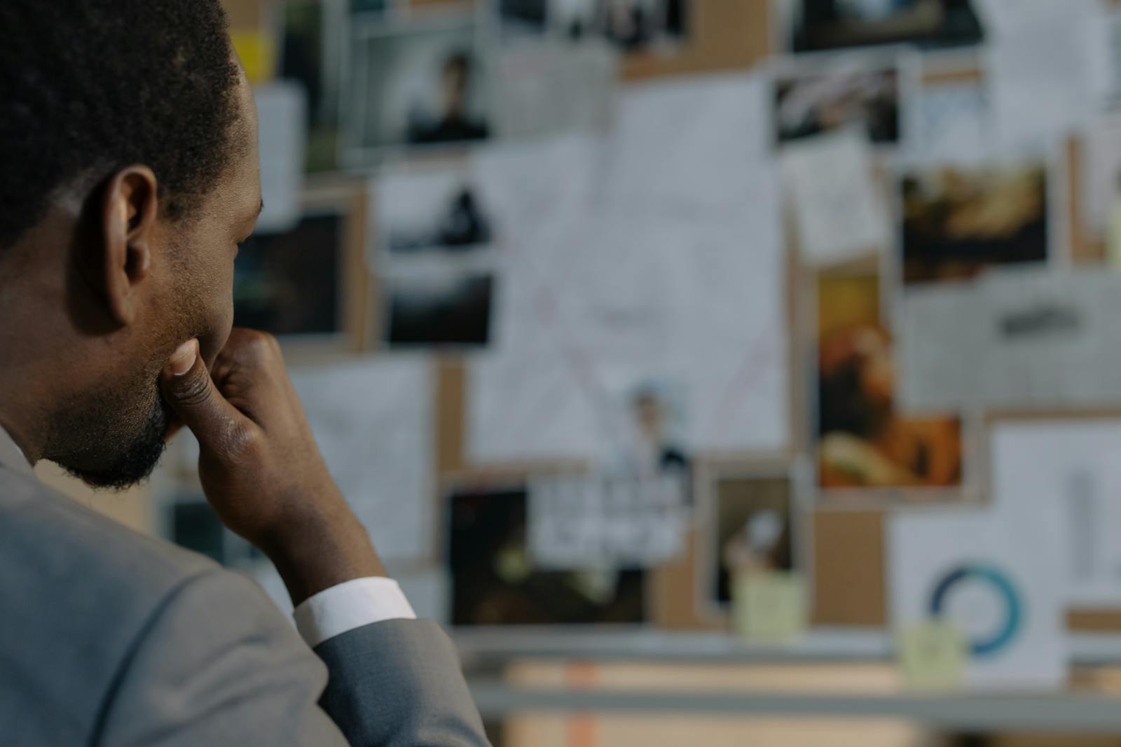 A detective in a suit examines an evidence board full of photos and documents.