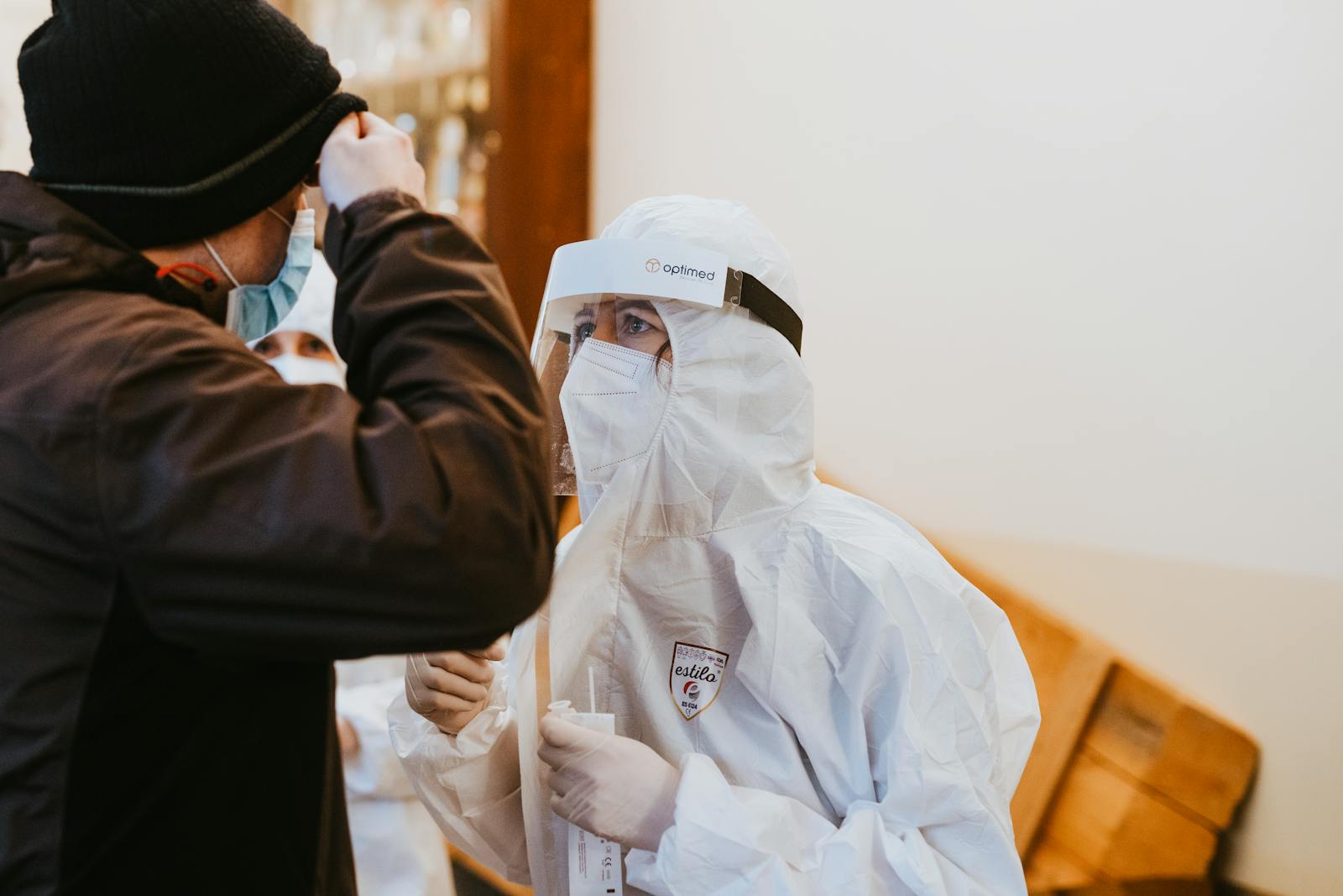 Healthcare professional wearing protective suit and face shield assisting a patient indoors.