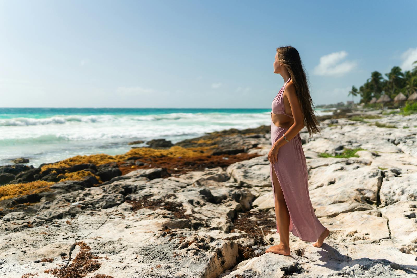 A woman in a flowing dress enjoys the scenic view of the rocky Tulum coastline under clear skies.