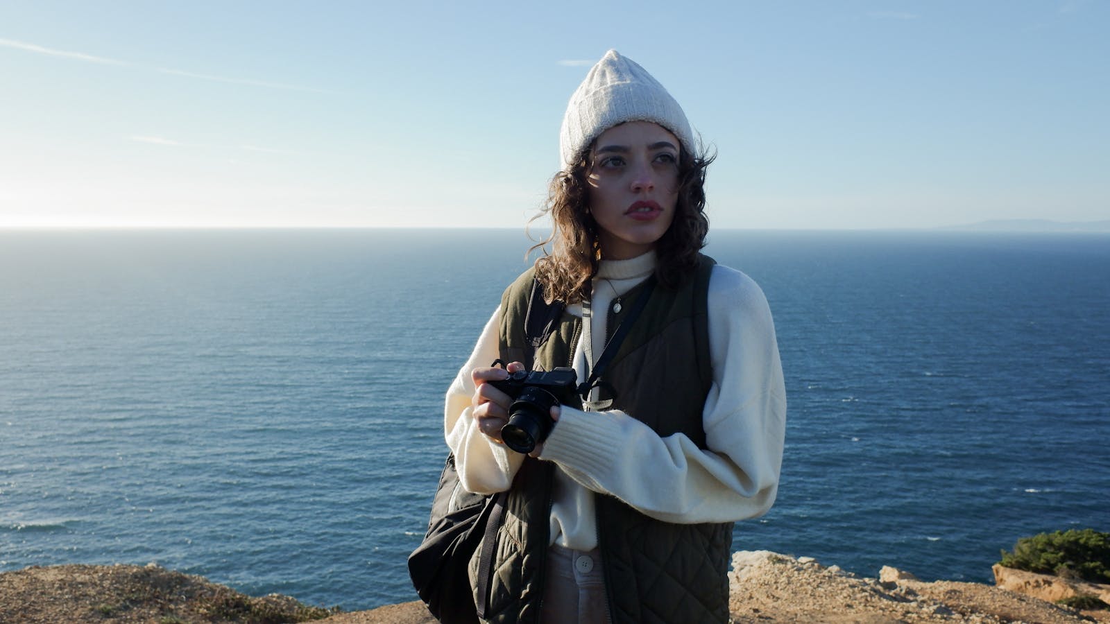 Woman standing on a cliff by the ocean holding a camera, wearing a beanie and puffer vest.