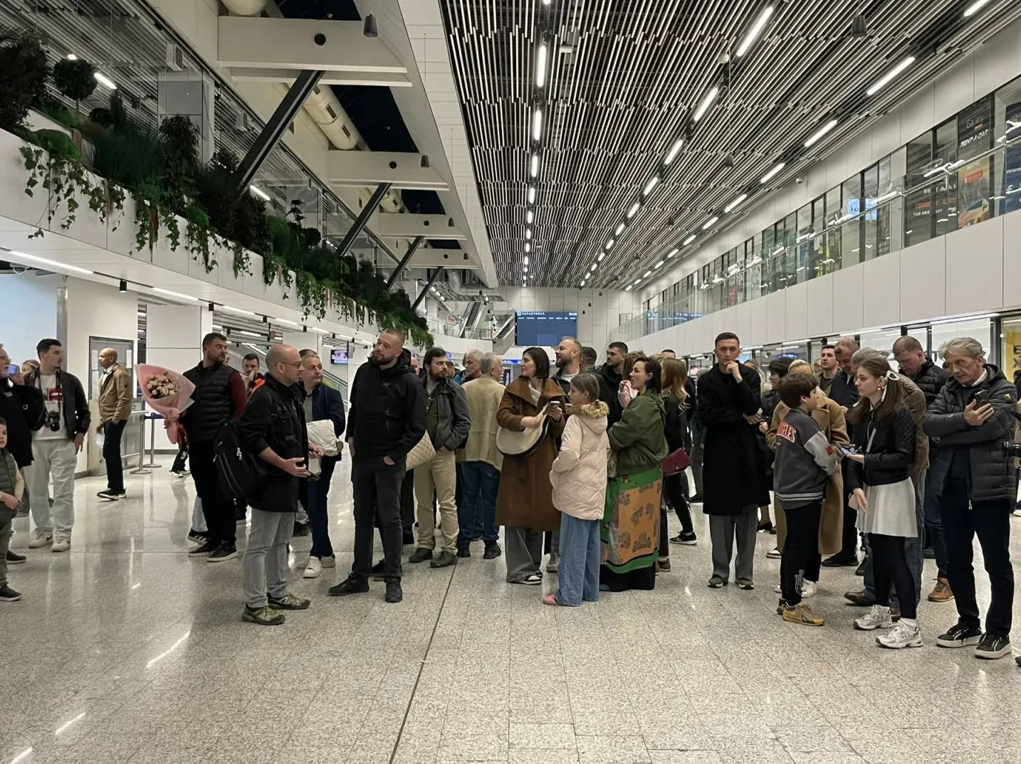 Grupa ljudi stoji u prostranom aerodromskom terminalu sa modernim osvjetljenjem i zelenilom na zidovima, neki drže prtljag, a jedan muškarac drži buket cvijeća.