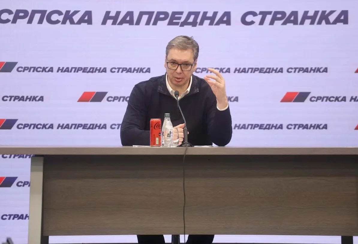 Aleksandar Vučić sjedi za stolom ispred pozadine s logom Srpske napredne stranke, držeći govor.