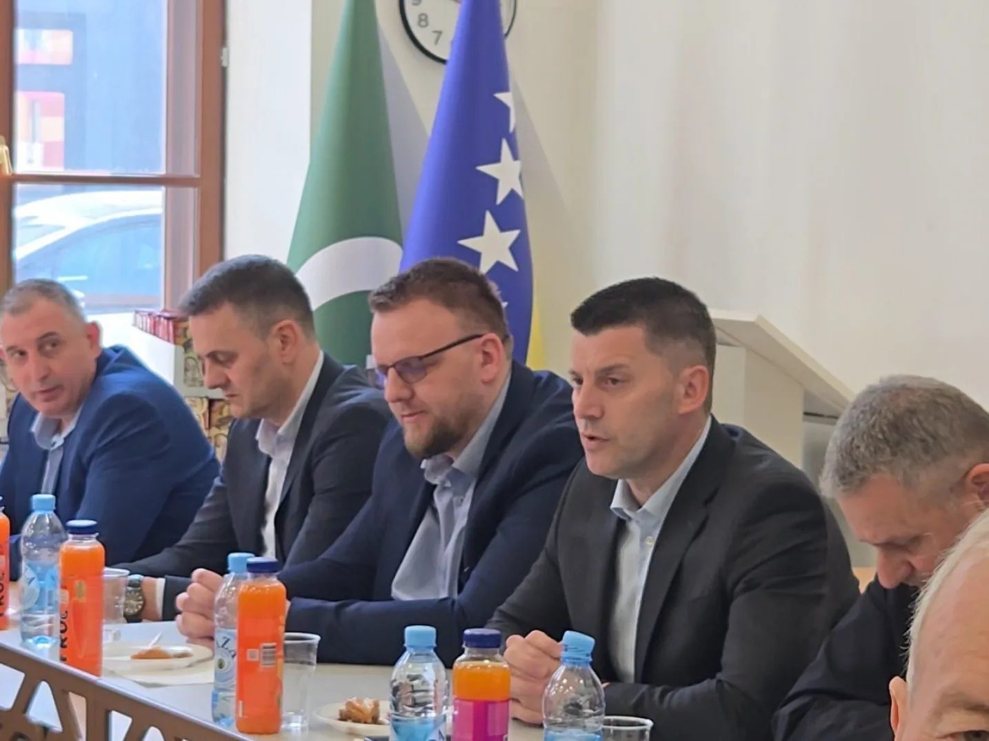 Grupa muškaraca sjedi za stolom, ispred njih su flaše vode i sokova. U pozadini su zastave Bosne i Hercegovine i Pakistana.