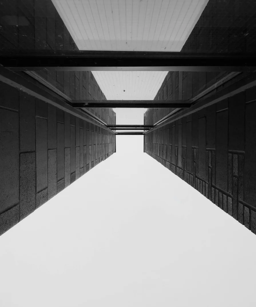 Crno-bijela fotografija koja prikazuje odraz arhitektonskog objekta u staklenom krovu, sa naglašenim detaljima betonskih fasada koje se sužavaju prema nebu.