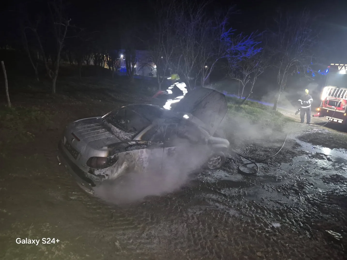 Izgorjeli automobil sa dimom koji izlazi, u noćnim uslovima, vatrogasci sa opremom pored vatrogasnog vozila.