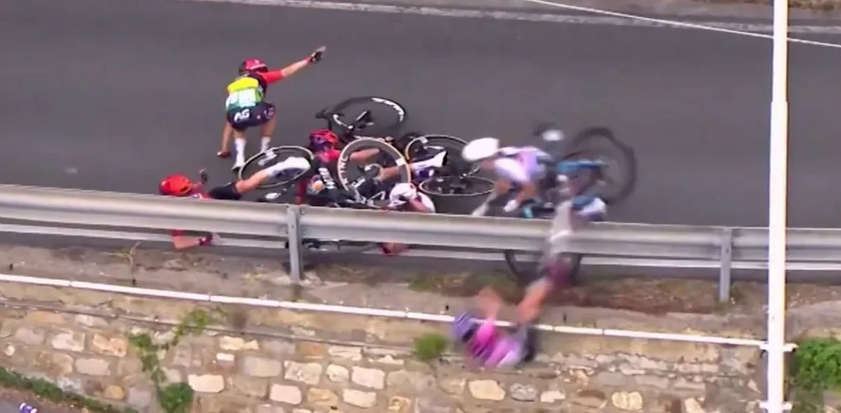 Više biciklista je palo na biciklističku trku, sa biciklima i opremom razbacanom po putu i pored zaštitne ograde.