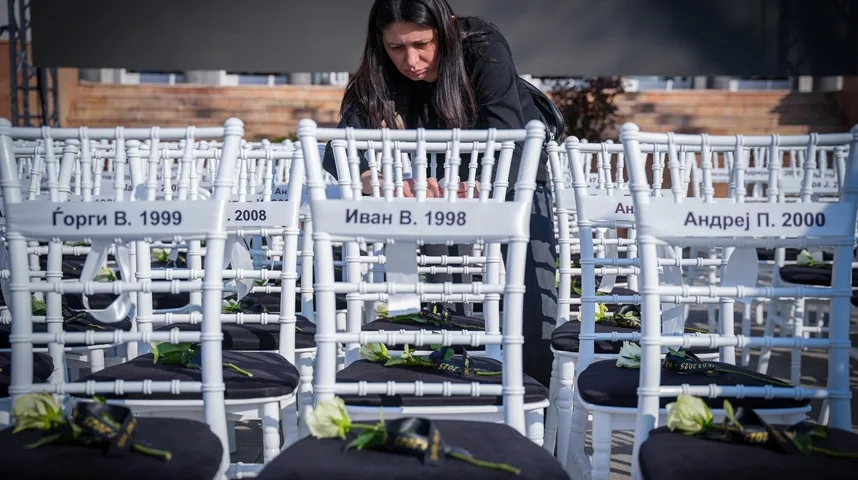 Blijedo osvijetljena fotografija prikazuje brojne bijele stolice poređane u redovima, koje su namijenjene za sjedenje. Na naslonima svake stolice nalazi se bijela traka ispisana crnim slovima, a na njoj je navedeno ime i godina rođenja osobe. Na crnim jastucima stolica leže bijele ruže, a neke od njih su ukrašene crnim vrpcama s natpisima.