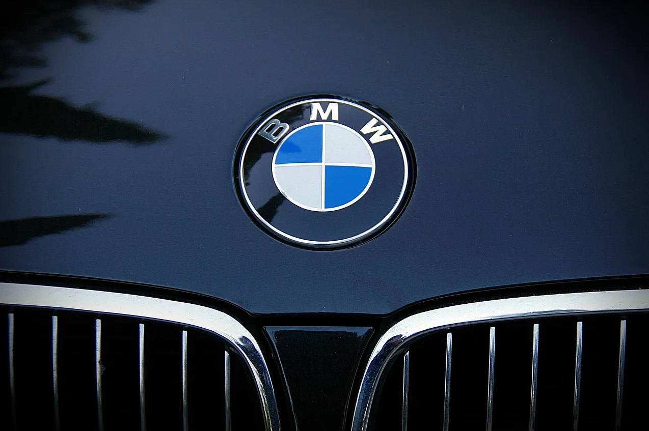 Bliski prikaz BMW-ovog logotipa smještenog na tamnoj haubi automobila, iznad prednje rešetke.