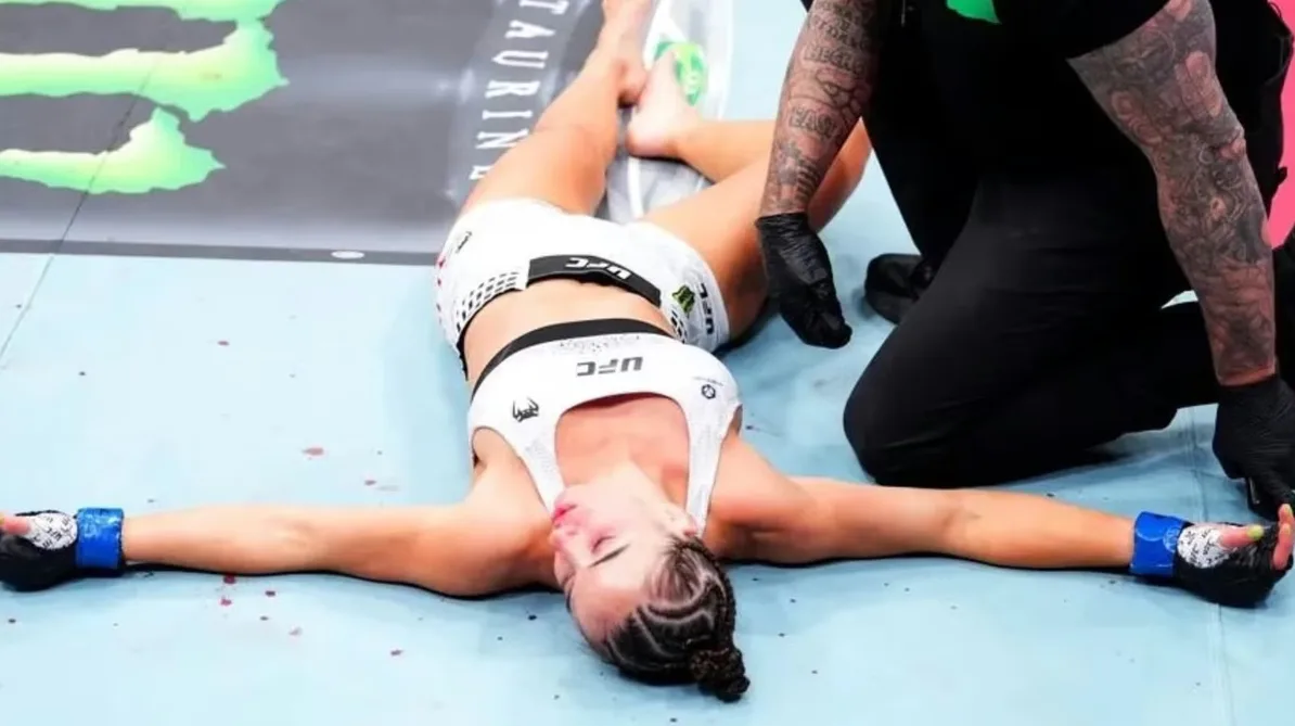 Ženska MMA borkinja leži na podu oktagona nakon borbe, s rukama raširenim u stranu i vidljivim povredama.