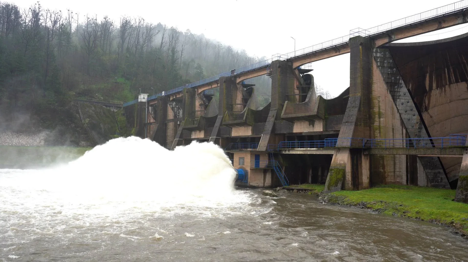 Brana sa snažnim izbacivanjem vode iz odvodnih cijevi uz maglovitu šumu u pozadini.