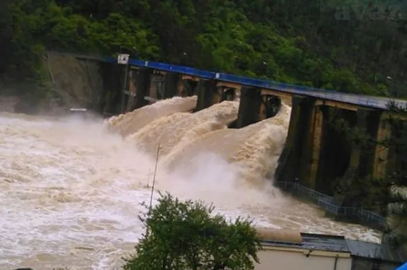Brana sa velikom količinom vode koja protiče i stvara pjenu, sa zelenilom u pozadini.