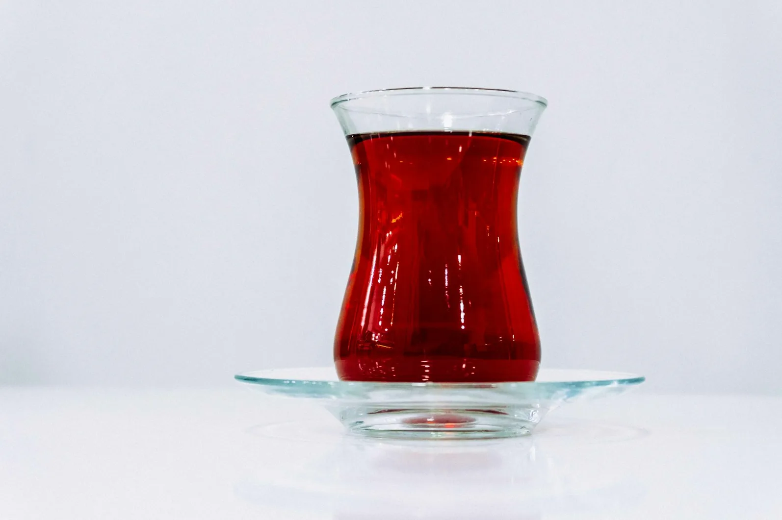 Staklo u obliku lale, tipično za posluživanje čaja, ispunjeno intenzivno crvenim napitkom. Staklo stoji na prozirnom tanjiriću, na bijeloj, glatkoj površini.