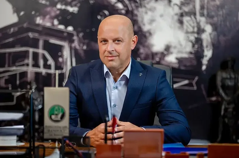 Ozbiljan muškarac ćelave glave u tamnoplavom odijelu i svijetloplavoj košulji sjedi za stolom u kancelariji.