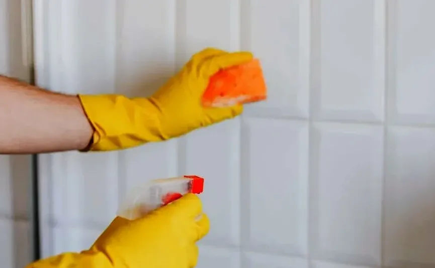 Osoba u žutim rukavicama čisti bijele pločice sunđerom i sprejem.