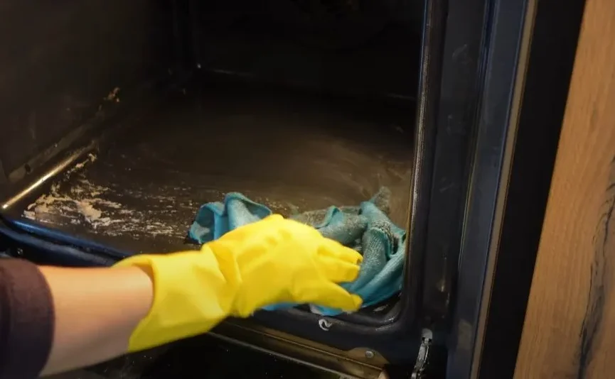 Ruka u žutoj zaštitnoj rukavici čisti unutrašnjost rerne plavom krpom.