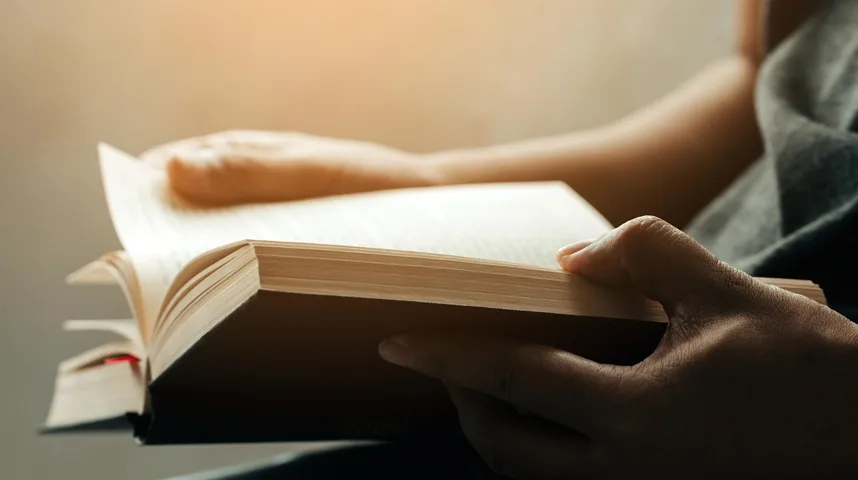 Osoba drži otvorenu knjigu i čita pri toplom svjetlu.