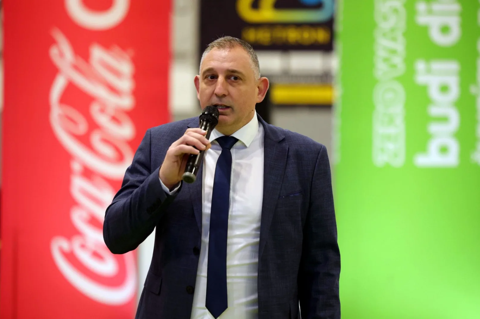 Muškarac u tamnoplavom odijelu i bijeloj košulji s tamnoplavom kravatom drži mikrofon i govori ispred Coca-Cola transparenta s lijeve strane i zelenog transparenta s natpisom "budi dio" s desne strane.