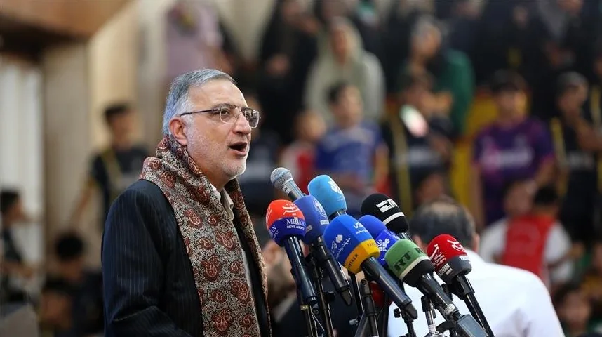 Političar ili javna ličnost, muškarac sa naočarima i šarenim šalom oko vrata, govori za govornicu sa više mikrofona ispred sebe. U pozadini se nazire mnoštvo ljudi.