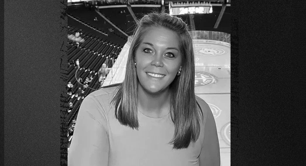 Crno-bijela fotografija nasmijane žene sa podignutom kosom, u ležernoj majici, sa pozadinom dvorane za hokej ili sličnog sportskog objekta.