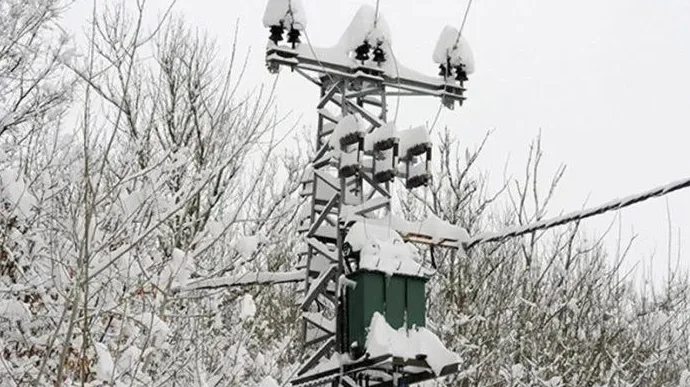 Stup dalekovoda prekriven snijegom sa transformatorom i izolatorima, okružen drvećem i granama pod snijegom.