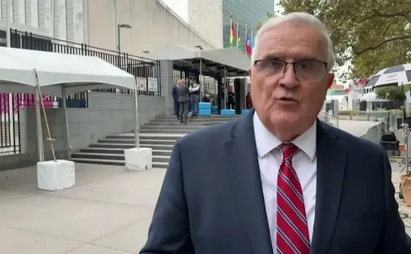 Diplomata u tamnom odijelu i crvenoj kravati sa bijelim prugama govori ispred sjedišta Ujedinjenih naroda.