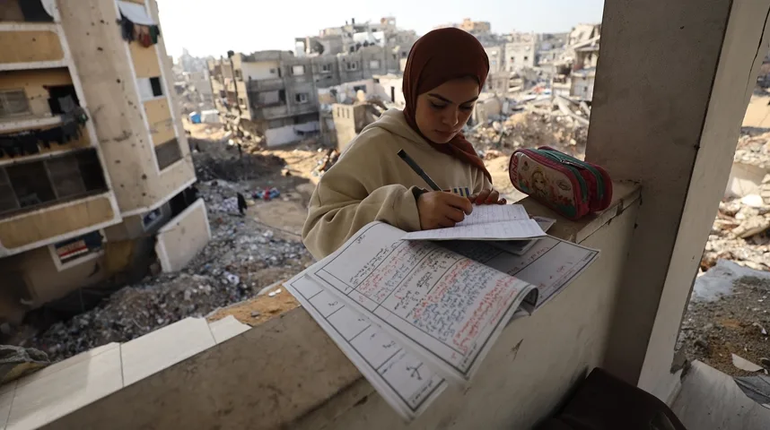 Mlada Palestinka u hidžabu piše u bilježnicu na balkonu uništene zgrade, dok se u pozadini vide ostaci drugih zgrada i ruševine.