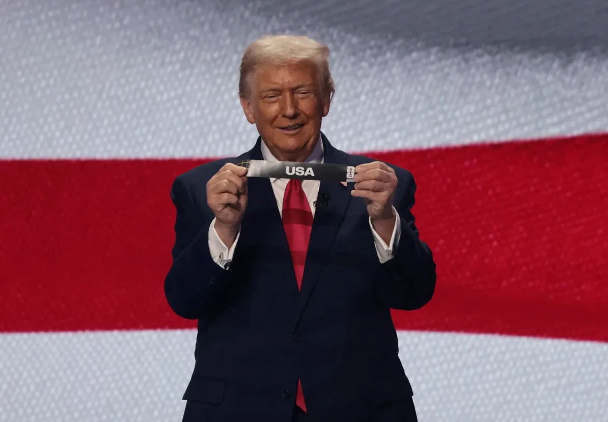 Donald Trump u tamnom odijelu i crvenoj kravati drži papirić s natpisom "USA" ispred pozadine sa crvenim i bijelim prugama.