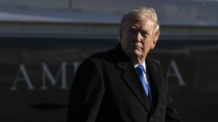 Donald Trump u crnom kaputu i plavoj kravati gleda u stranu, sa natpisom "AMERICA" na pozadini aviona.