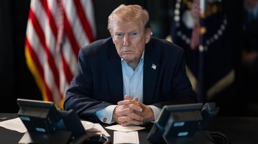 Donald Trump sjedi za stolom s rukama spojenim ispred sebe, ispred zastave Sjedinjenih Američkih Država.