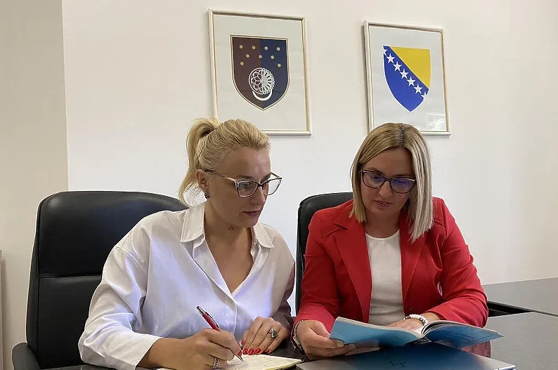 Dvije žene sede za stolom u kancelariji, jedna piše u bilježnicu, a druga čita dokument, sa državnim grbom Bosne i Hercegovine i zastavom na zidu u pozadini.