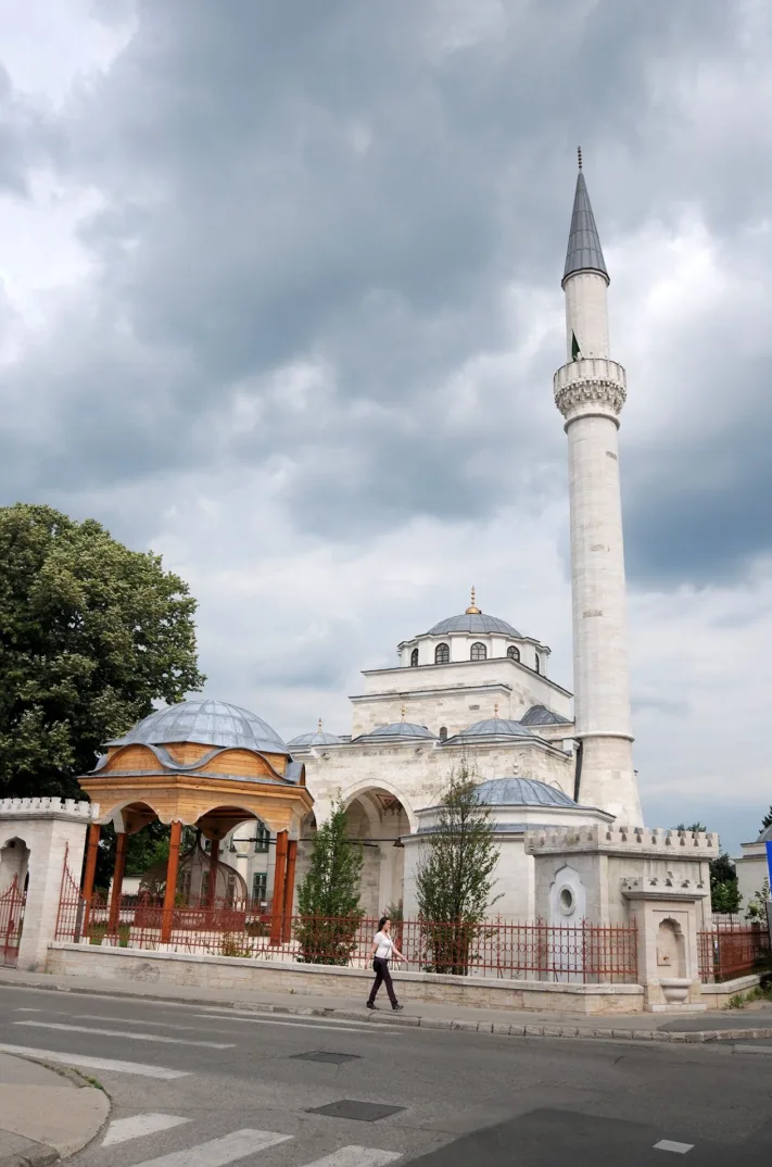 Bijela džamija sa sivim kupolama i visokim, uskim minaretom na oblačno nebo.