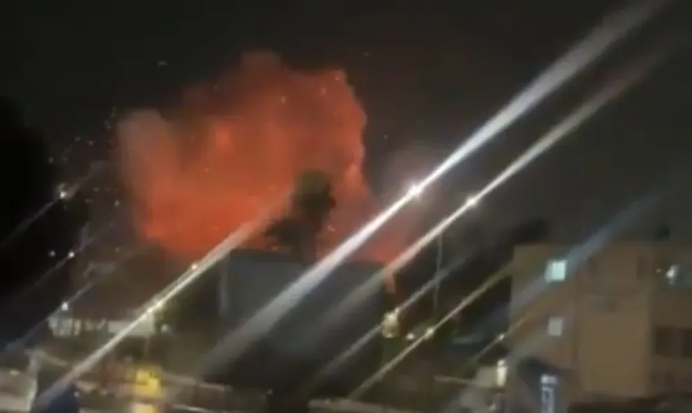 Noćni snimak eksplozije sa jarkim narandžastim plamenom i iskricama iznad zgrada, uz svjetlosne zrake.