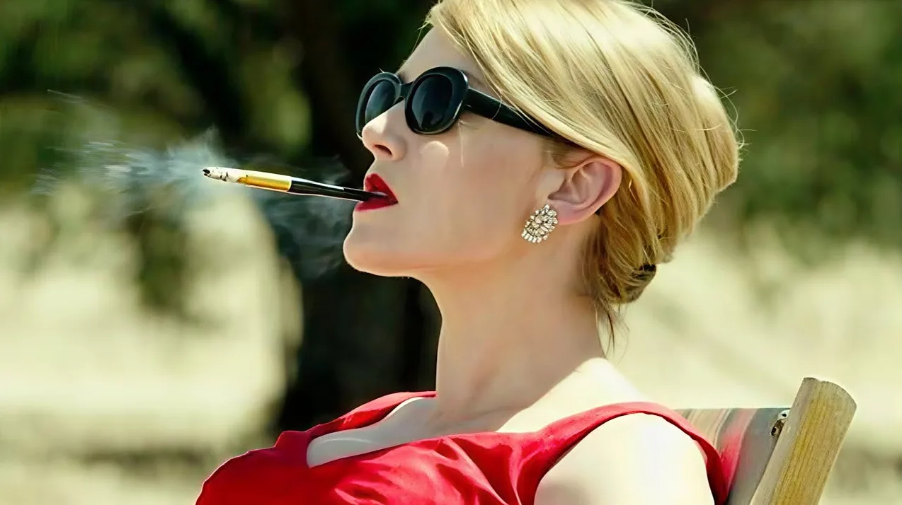 Elegantna plavuša u crvenoj haljini i velikim sunčanim naočarima puši cigaretu iz usnika, sa dimom koji izlazi iz nje.
