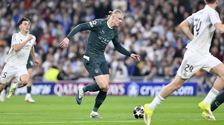 Fudbaler Erling Haaland u tamnozelenom dresu Mančester sitija trči sa loptom na terenu, dok ga prate igrači Real Madrida.