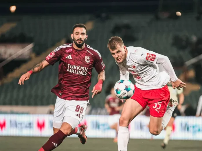 Фудбалер у бијелом дресу са броjем 35 трчи са лоптом, док га у потери прати фудбалер у бордо дресу.