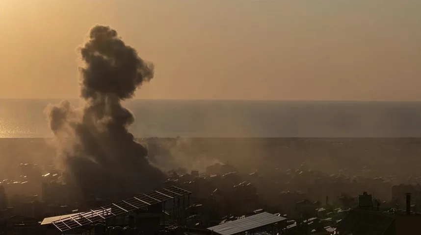 Veliki stub tamnog dima diže se sa urbanog područja prema nebu, iznad kojeg se pruža horizont mora i blijedo narandžasto nebo.