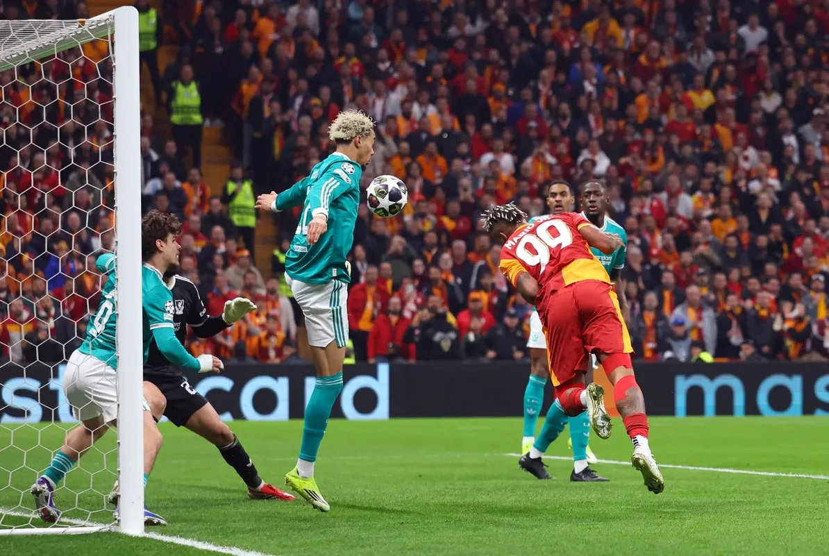 Fudbalski igrač u zelenom dresu skače i prima loptu glavom, dok ga drugi igrač u crvenom dresu sa brojem 99 pokušava blokirati, a golman i drugi igrači su u blizini.