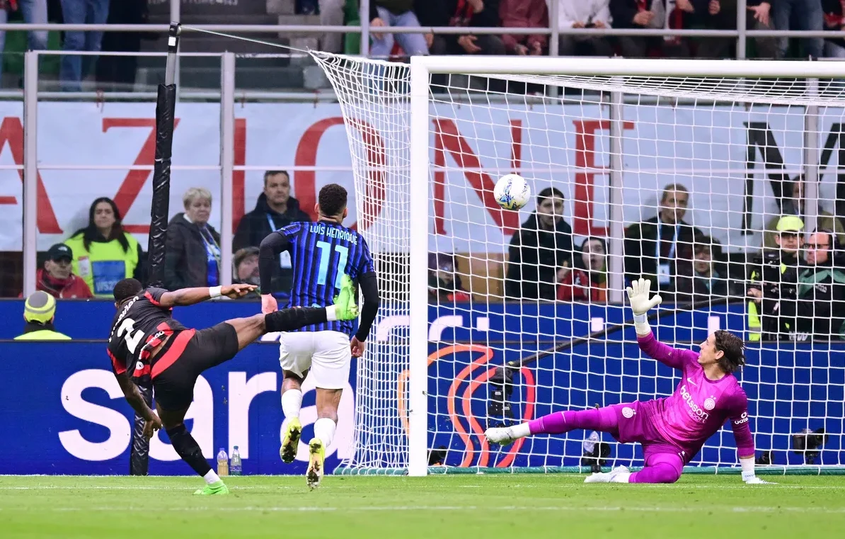 Fudbaler u crno-crvenom dresu šutira prema golu, a golman u ljubičastom dresu se baca kako bi odbranio udarac. Lopta se nalazi u vazduhu blizu gola.