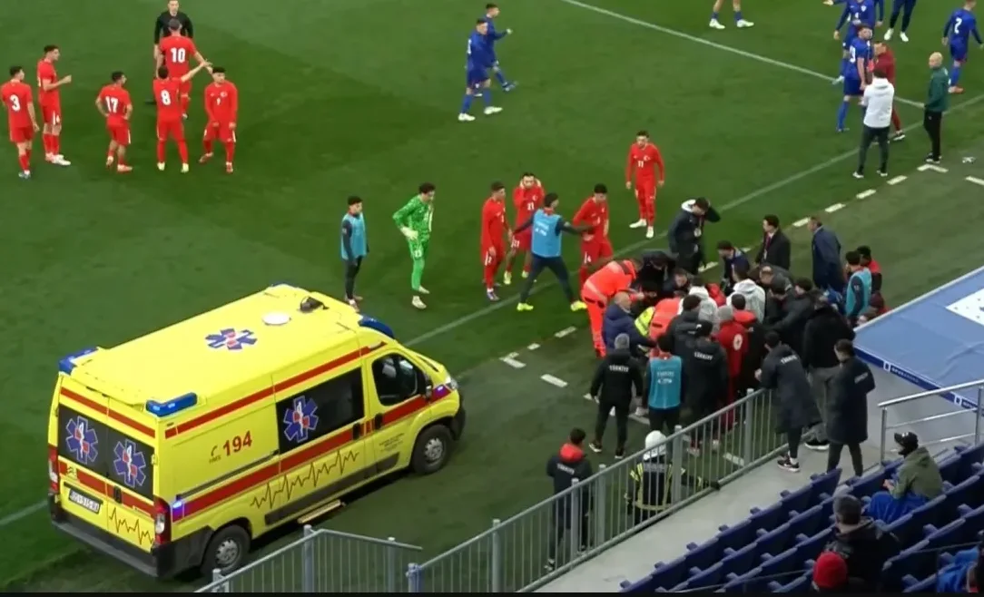 Fudbalski teren sa igračima u crvenim dresovima, medicinskim osobljem, ekipom hitne pomoći i vozilom hitne pomoći.