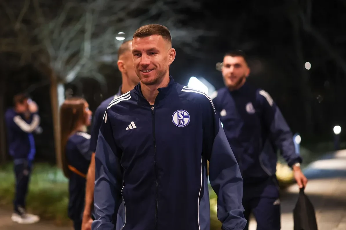 Fudbaler tima Schalke 04, odjeven u tamnoplavu Adidas trenerku s amblemom kluba, osmjehuje se dok hodaju noću. U pozadini se naziru ostali članovi tima.