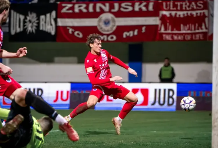 Fudbaler u crvenom dresu, sa brojem 4 na leđima, izvodi udarac prema golu. Lopta je u vazduhu ispred njega, a teren je prekriven vještačkom travom. U pozadini se vide navijači i transparenti kluba.