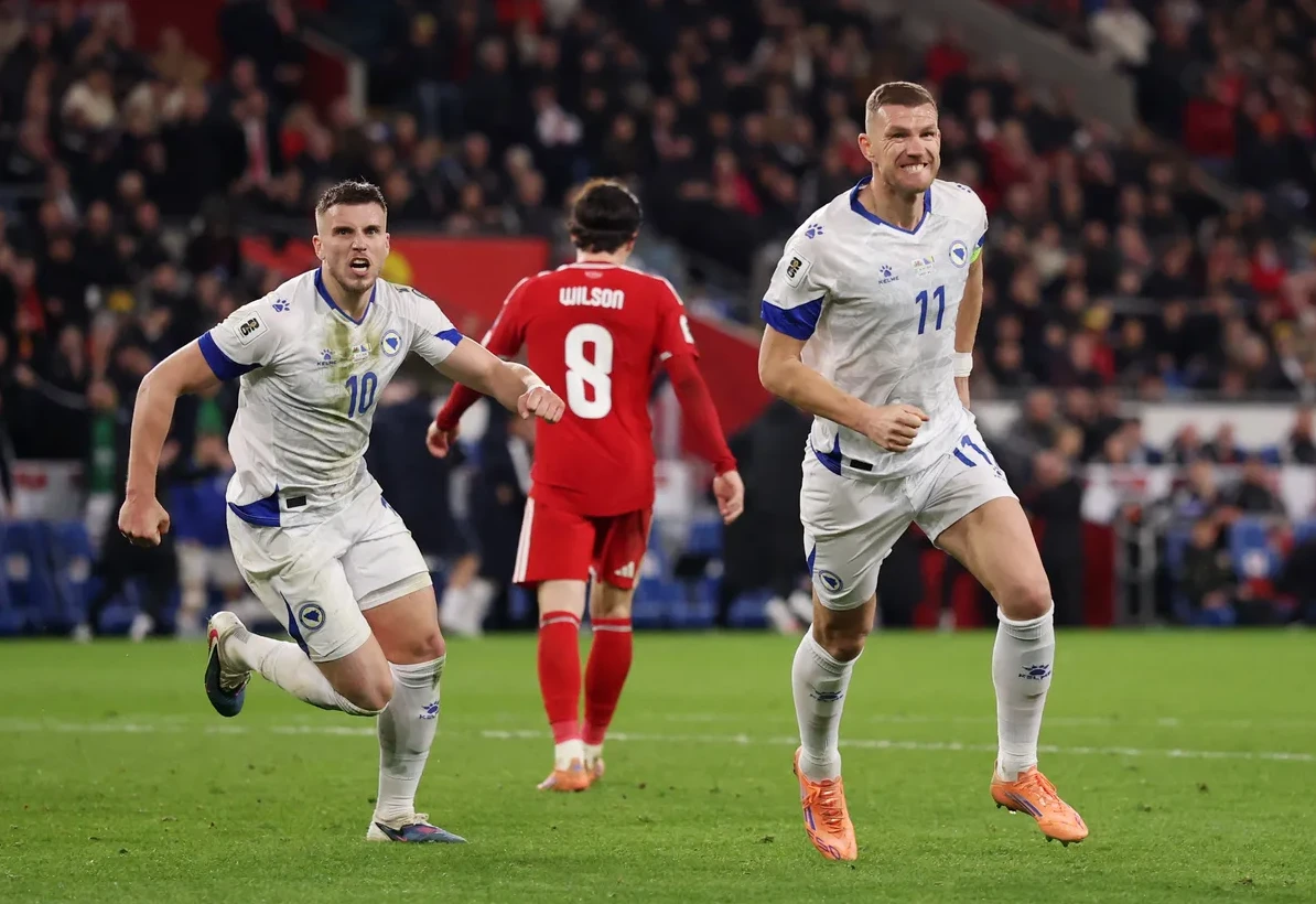 Dva fudbalera Bosne i Hercegovine u bijelim dresovima trče po zelenom travnjaku fudbalskog terena.