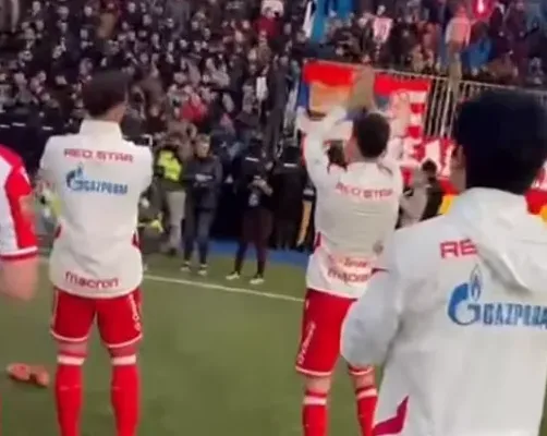 Fudbaleri Crvene zvezde u bijelim dresovima sa crvenim detaljima, na čijim se leđima vide logotipi "GAZPROM" i "Macron", pozdravljaju navijače koji stoje na tribinama.
