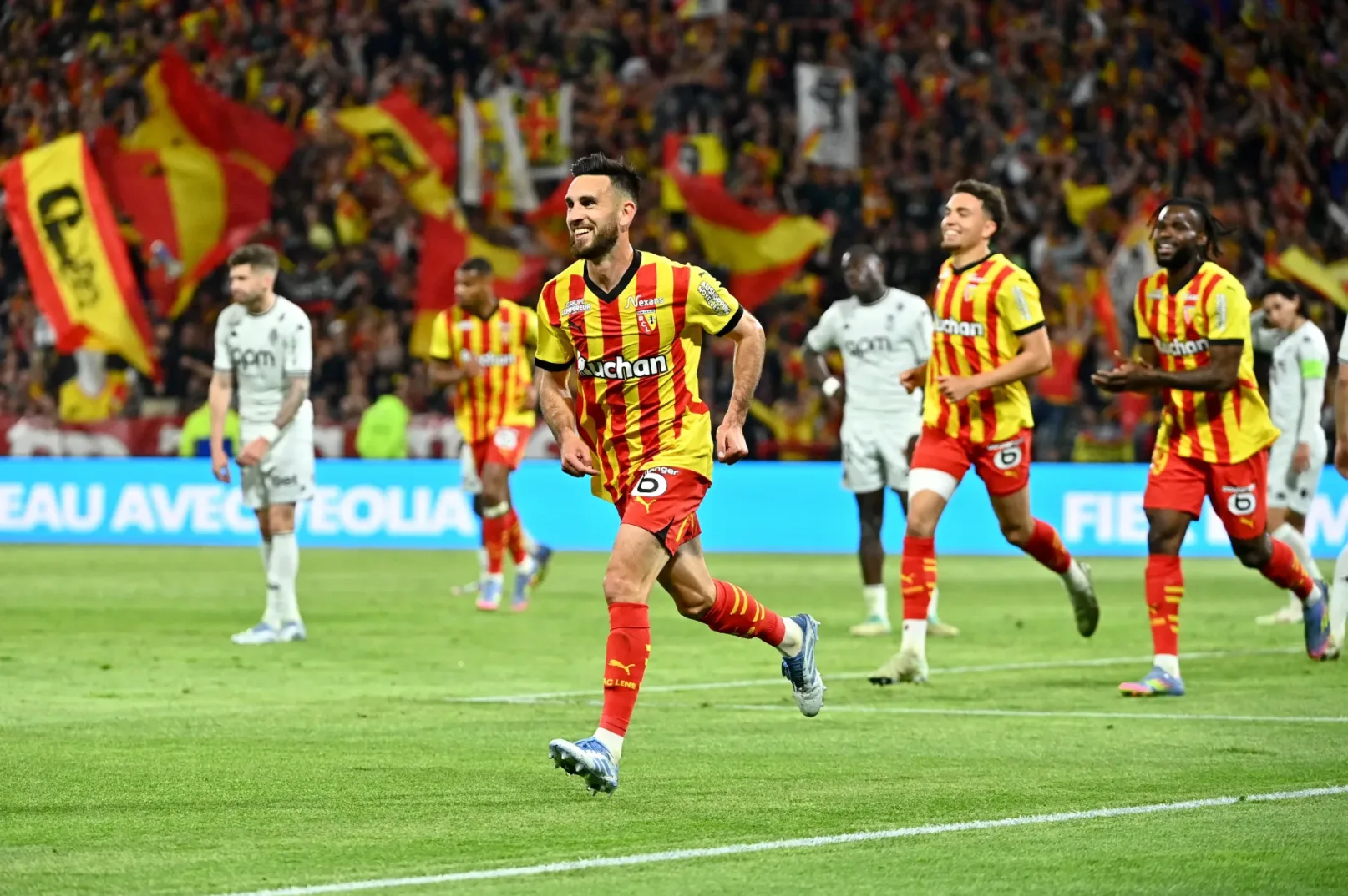 Fudbaler RC Lensa, sa brojem 28 na leđima, trči po terenu sa osmijehom na licu, obučen u crveno-zlatni dres sa natpisom 'Auchan' i kratkim rukavima. Njegovi saigrači, također u sličnim dresovima, slave gol iza njega. U pozadini se vidi puna tribina navijača sa crveno-žutim zastavama. Na terenu su prisutni i igrači protivničke ekipe u bijelim dresovima.