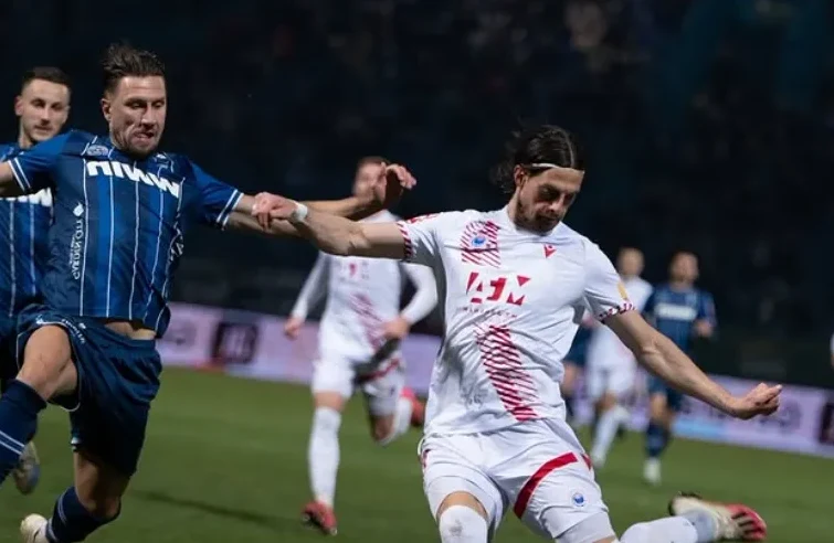 Fudbaler u bijelom dresu sa crvenim detaljima izvodi loptu, dok ga ometa protivnički igrač u plavom dresu.